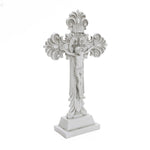 White ornate cross featuring a depiction of Jesus crucified, ideal for religious occasions. Detailed designs grace the ends, with inscriptions visible above Jesus. This symbol of Christian faith stands proudly on a rectangular base.