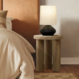 An oak finish side table with a black base, topped by a cream-shaded lamp, complements the bed adorned with beige blankets and pillows.