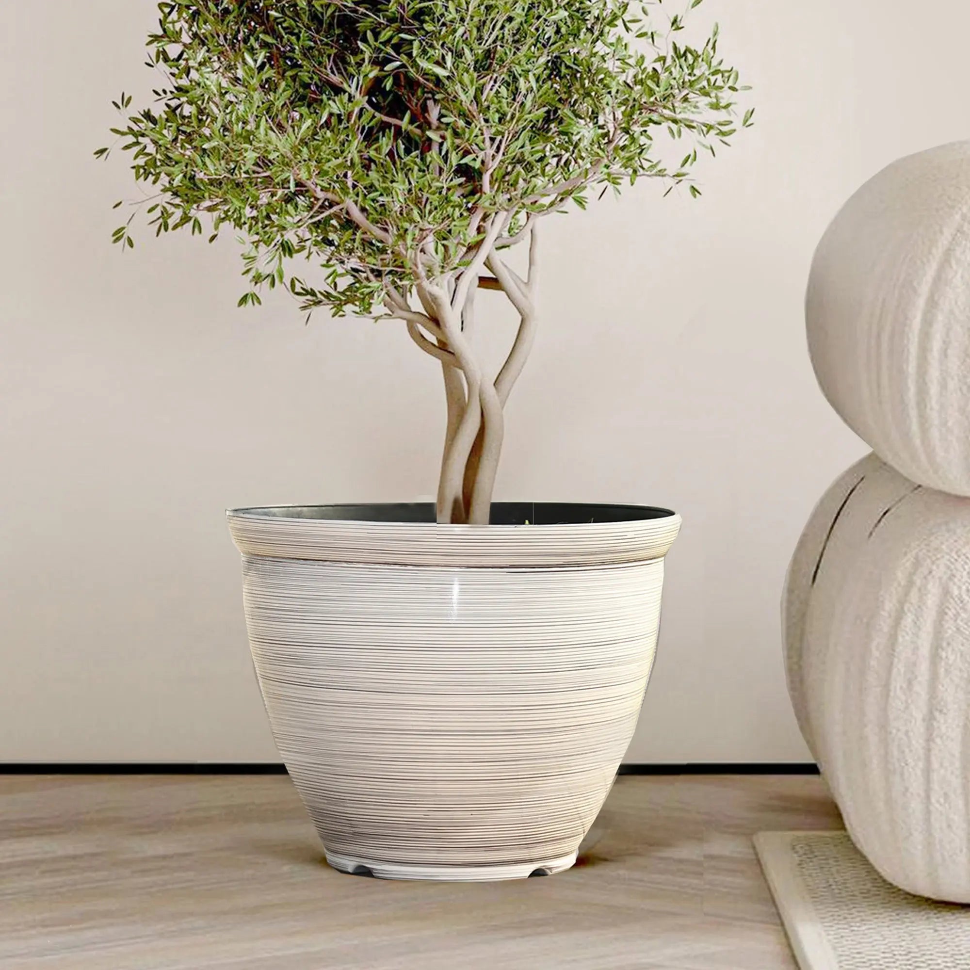 A contemporary planter featuring a potted tree in a light-colored ceramic design sits next to a round, textured cushion on the wooden floor, blending modern elegance with natural charm.