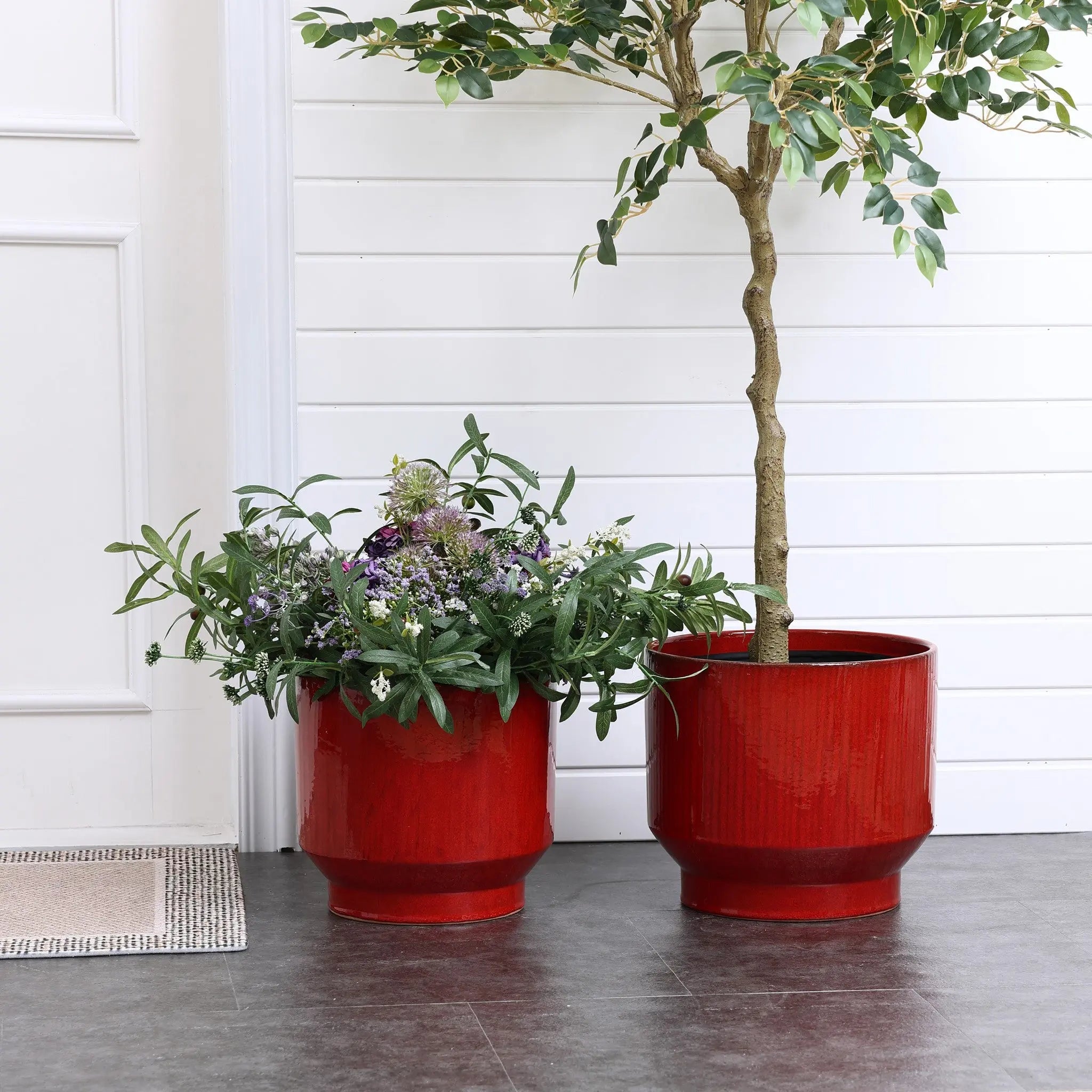 Two burgundy red glazed round ceramic planters from LuxenHome are on a dark floor. The left holds assorted green foliage with small purple and white flowers, while the right has a small tree. Both feature vertical stripes, set against a white wall and door backdrop with a partially visible doormat.
