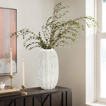 A LuxenHome Modern geometric pattern resin tall vase, 13.5 h in white holds green leafy branches on a dark wooden cabinet. Nearby are two tall white candles in gold holders. A large window provides natural light, with part of an abstract painting visible on the wall.