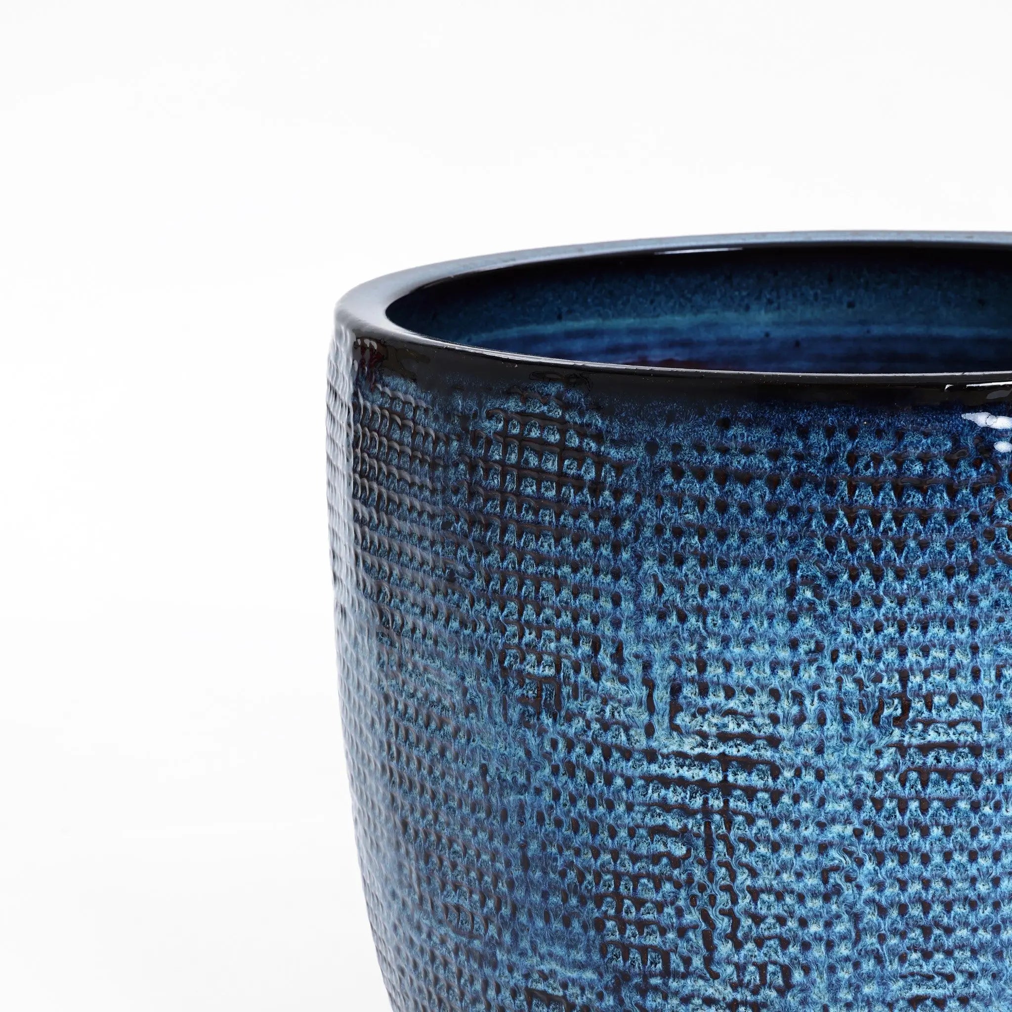 A close-up showcases the LuxenHome Ocean blue glazed round ceramic planter, 13.98 h, featuring a textured surface with a rich, glossy blue finish. Its intricately designed top edge stands out against a plain white background, enhanced by a subtle gradient for an elegant look.