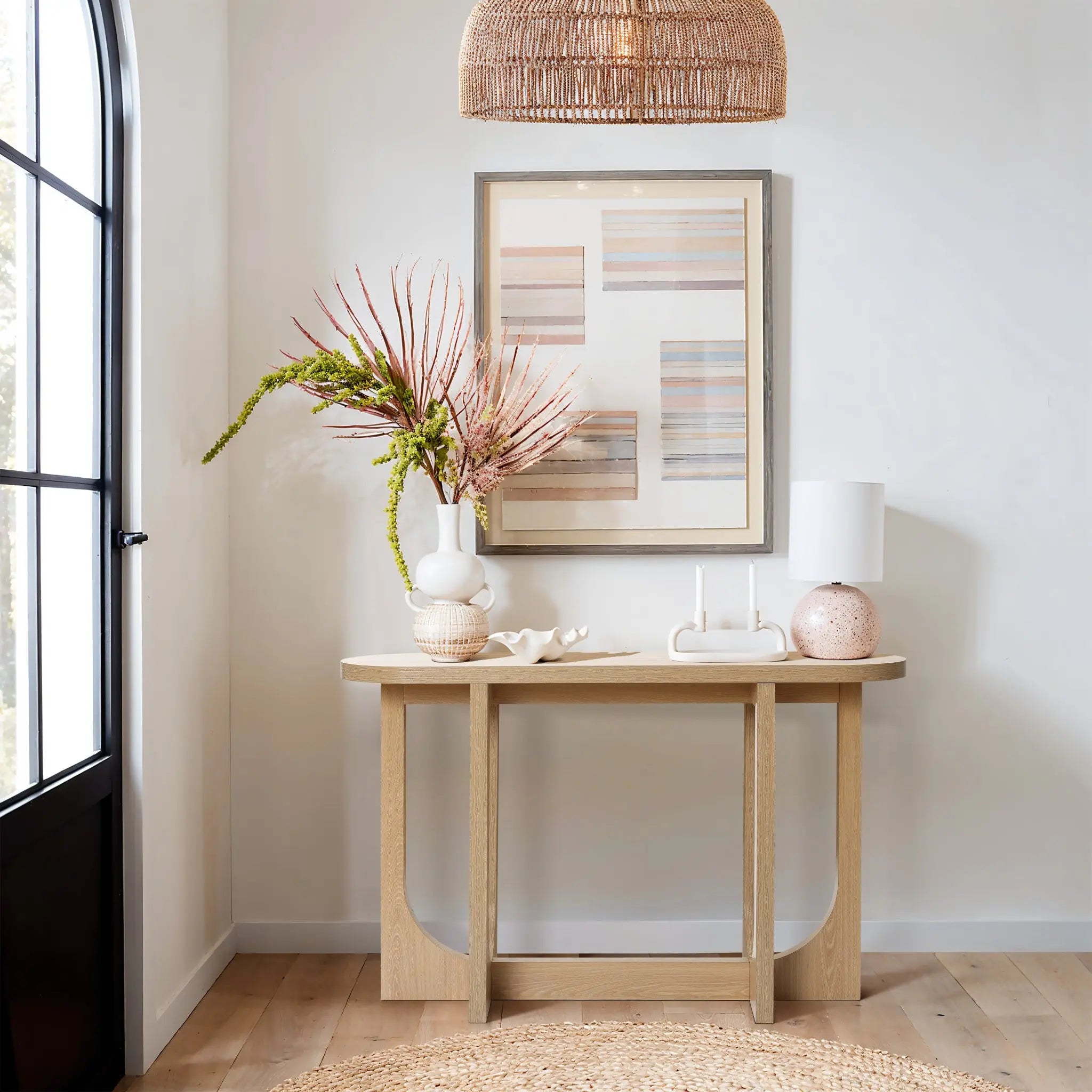 A stylish entryway features the Lorean natural finish contemporary console table by LuxenHome, adorned with vases and decor. An abstract framed artwork hangs above. The area is lit by a woven pendant light, with sunlight streaming through a tall window.