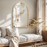 A cozy living room features a light beige sofa and a 34-inch Arch Mirror with scalloped edge framing. A vase of flowers complements the wooden decor, while sunlight streams through the window, casting shadows on the wall.