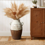 The LuxenHome rustic cream and brown terracotta urn vase with handles, 18.5 h, holds fluffy beige pampas grass for a touch of rustic elegance. Its placed on a wooden floor beside a cabinet adorned with a small plant and candle against a white paneled wall.