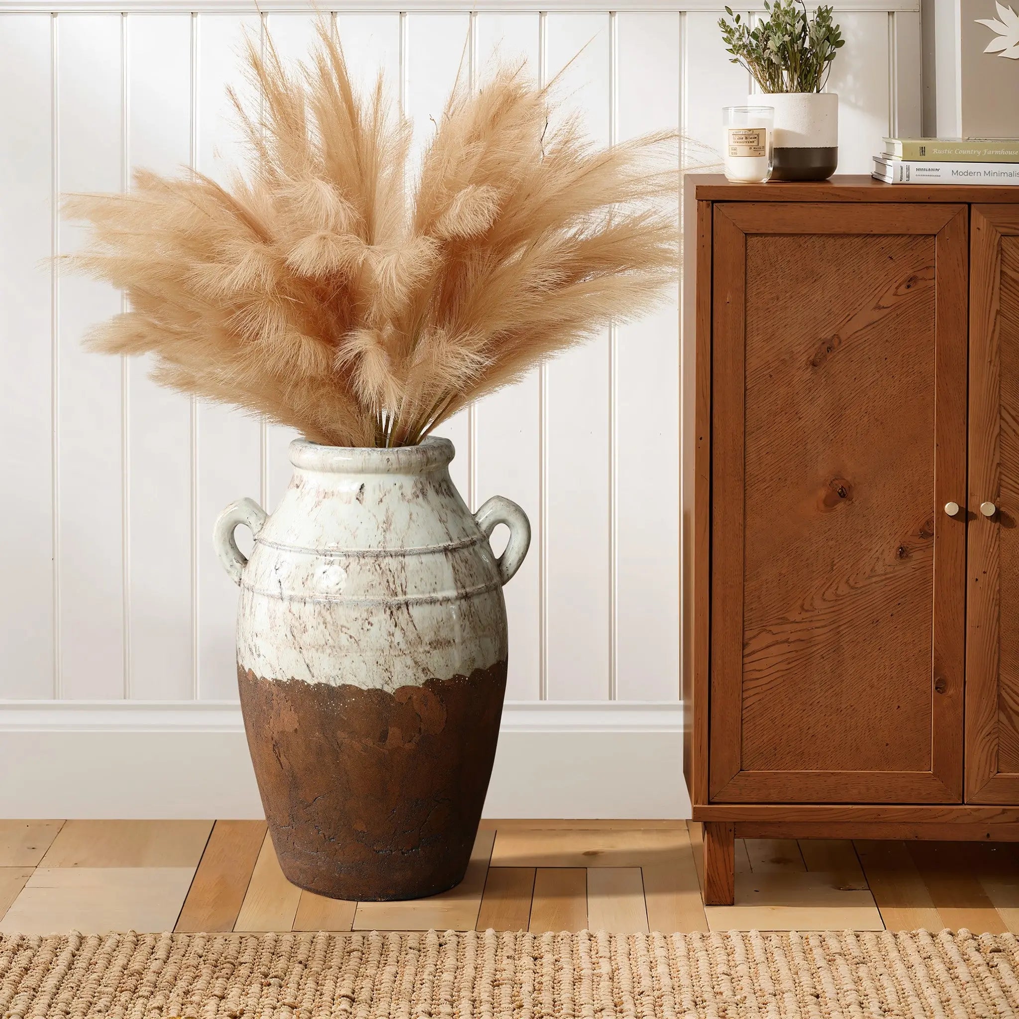 The LuxenHome rustic cream and brown terracotta urn vase with handles, 18.5 h, holds fluffy beige pampas grass for a touch of rustic elegance. Its placed on a wooden floor beside a cabinet adorned with a small plant and candle against a white paneled wall.