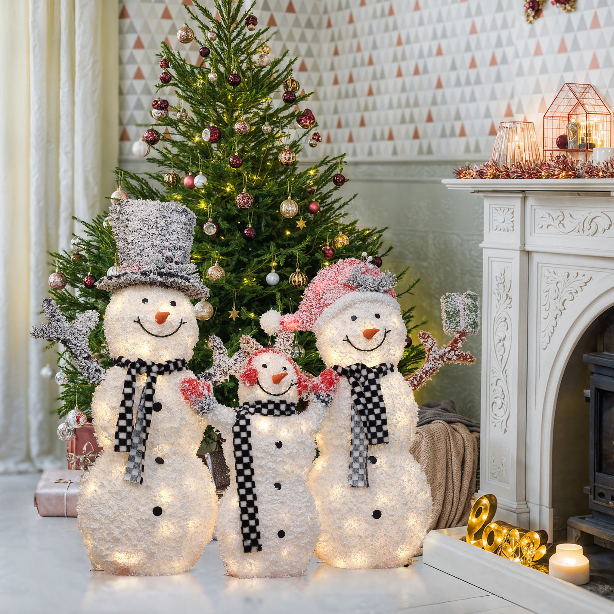 Three illuminated lighted snowman decorations with black-and-white scarves stand in front of a decorated Christmas tree beside a fireplace in a festive living room.