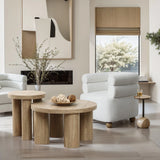 Modern living room featuring light wood tables, a white armchair, and wall art. A round side table with a drink complements the decor, while a large window reveals a tree outside. A vase with branches adds an elegant touch beside the oak finish side table.