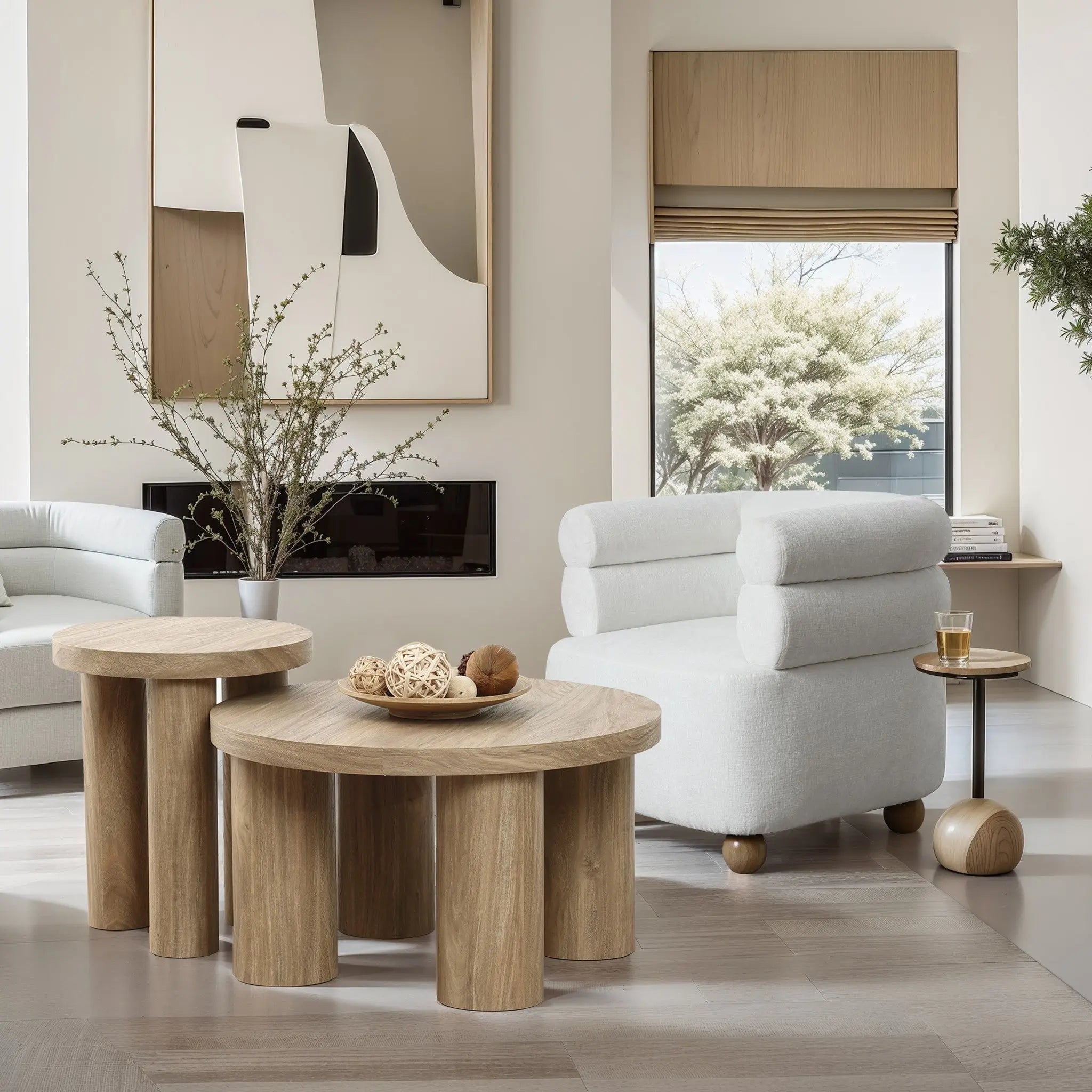 Modern living room featuring light wood tables, a white armchair, and wall art. A round side table with a drink complements the decor, while a large window reveals a tree outside. A vase with branches adds an elegant touch beside the oak finish side table.