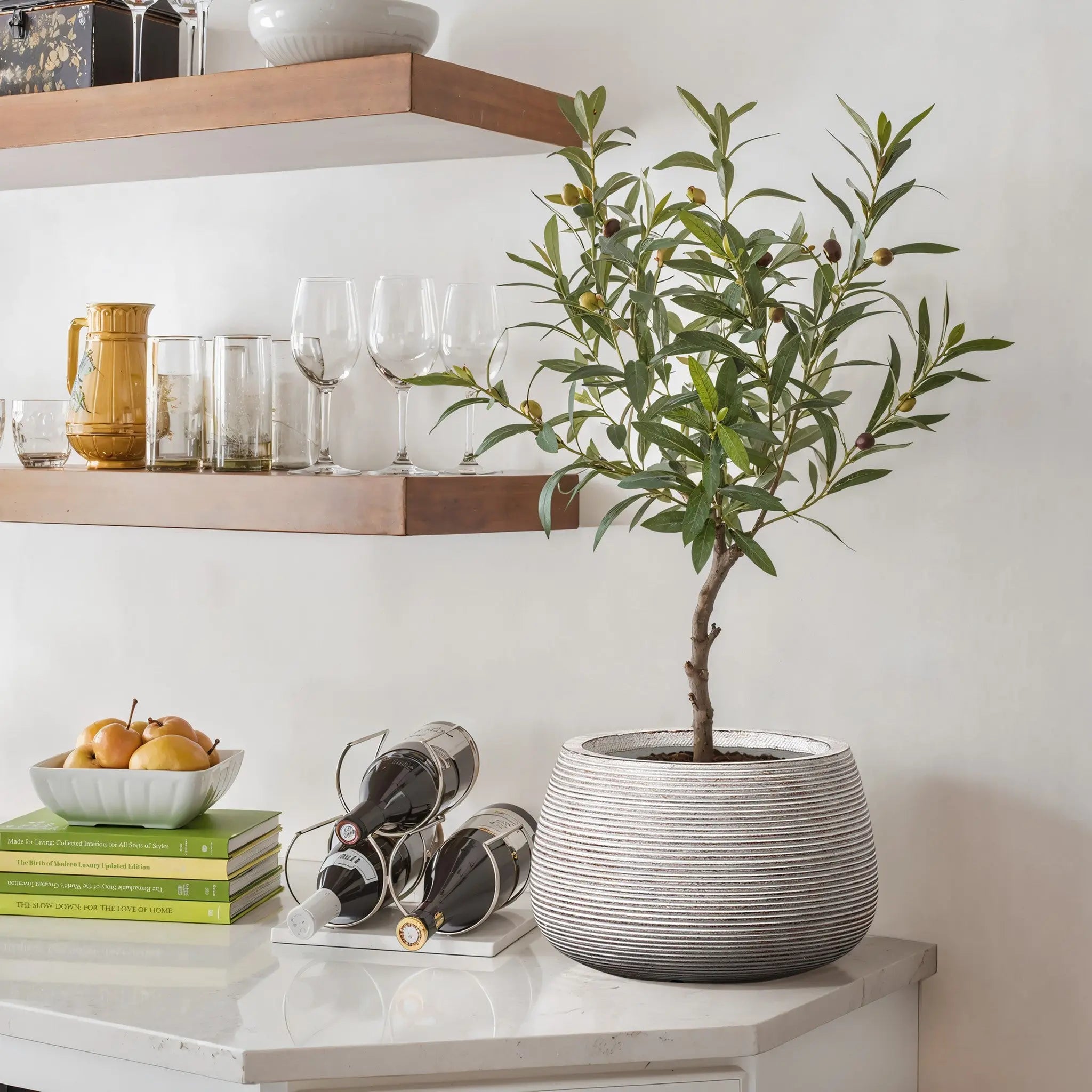 Sleek planter on a marble countertop with stacked books, a bowl of apples, and wine bottles. Wooden shelves above hold glassware in a minimalist design.