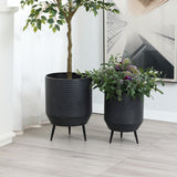 Two black planters on legs, boasting a distressed black finish, hold vibrant life; one cradles a tall leafy plant, while the other nurtures assorted greenery and purple flowers. They elegantly stand on a light wooden floor against a white wall adorned with a framed picture.