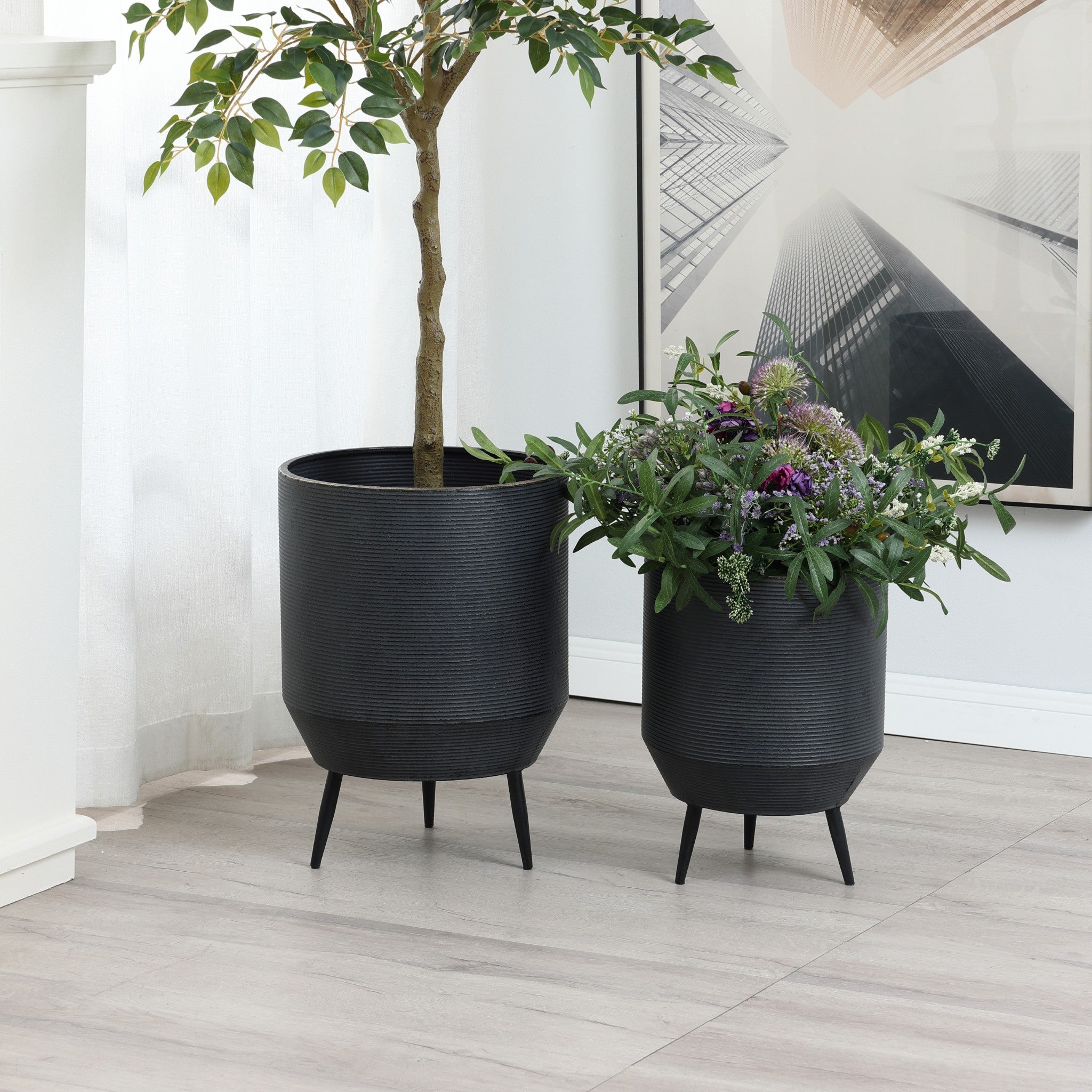 Two black planters on legs, boasting a distressed black finish, hold vibrant life; one cradles a tall leafy plant, while the other nurtures assorted greenery and purple flowers. They elegantly stand on a light wooden floor against a white wall adorned with a framed picture.