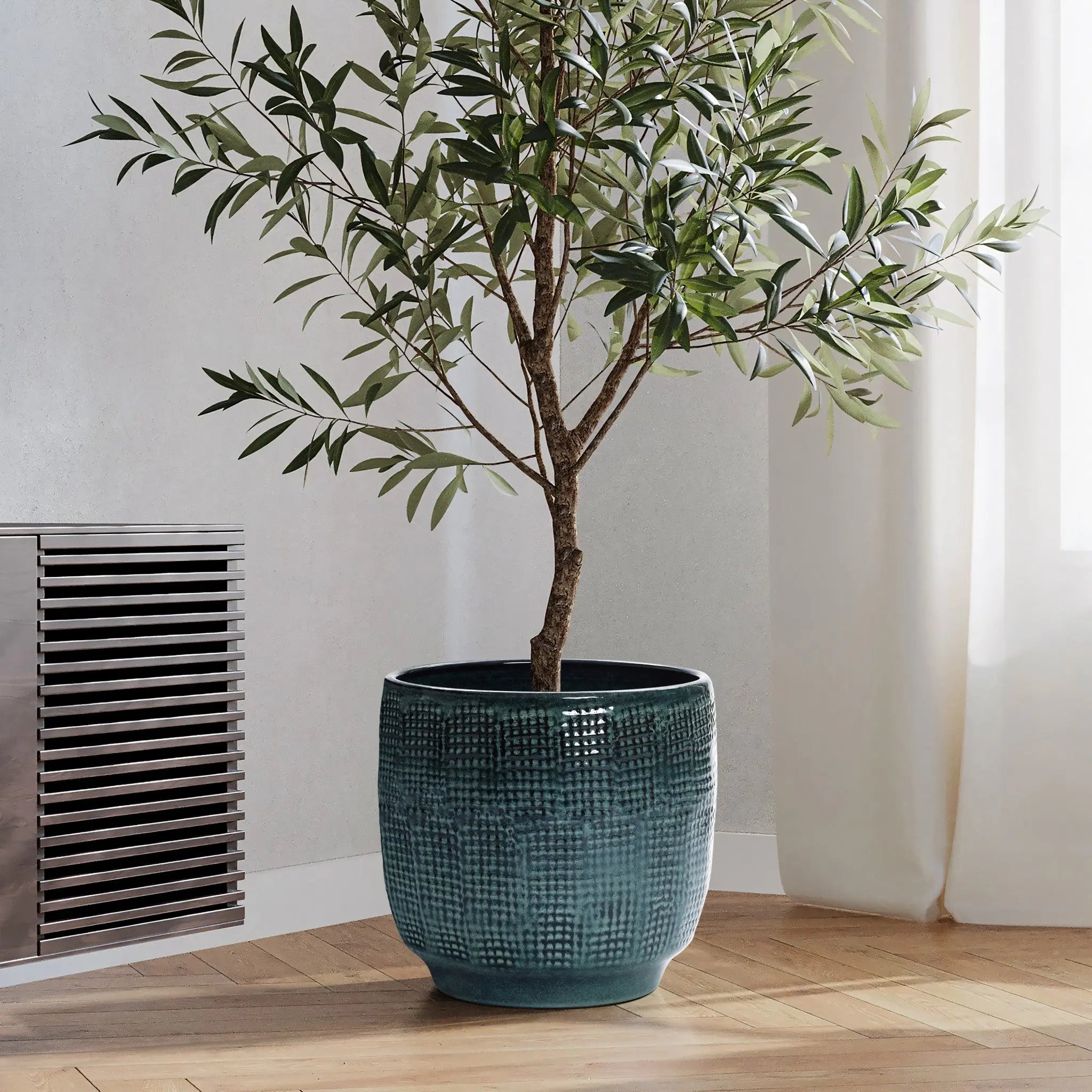 A tall indoor plant with slender branches and green leaves sits in a LuxenHome Marina gray glazed round ceramic planter, 11.22 h, on a wooden floor near a white curtain and metal wall vent, softly illuminated by natural light.