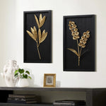 Two black-framed wall art pieces featuring golden leaves and flower bouquets hang above a shelf with a white vase, a small plant, and books, adding a touch of elegance to the interior design.