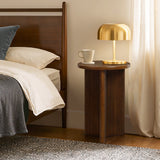 A mid-century modern side table with a round tabletop holds a golden lamp and a white mug beside a bed adorned with white linens. The room features a woven rug and curtains, adding an elegant touch. Crafted from high-quality MDF wood, it exudes both style and durability.