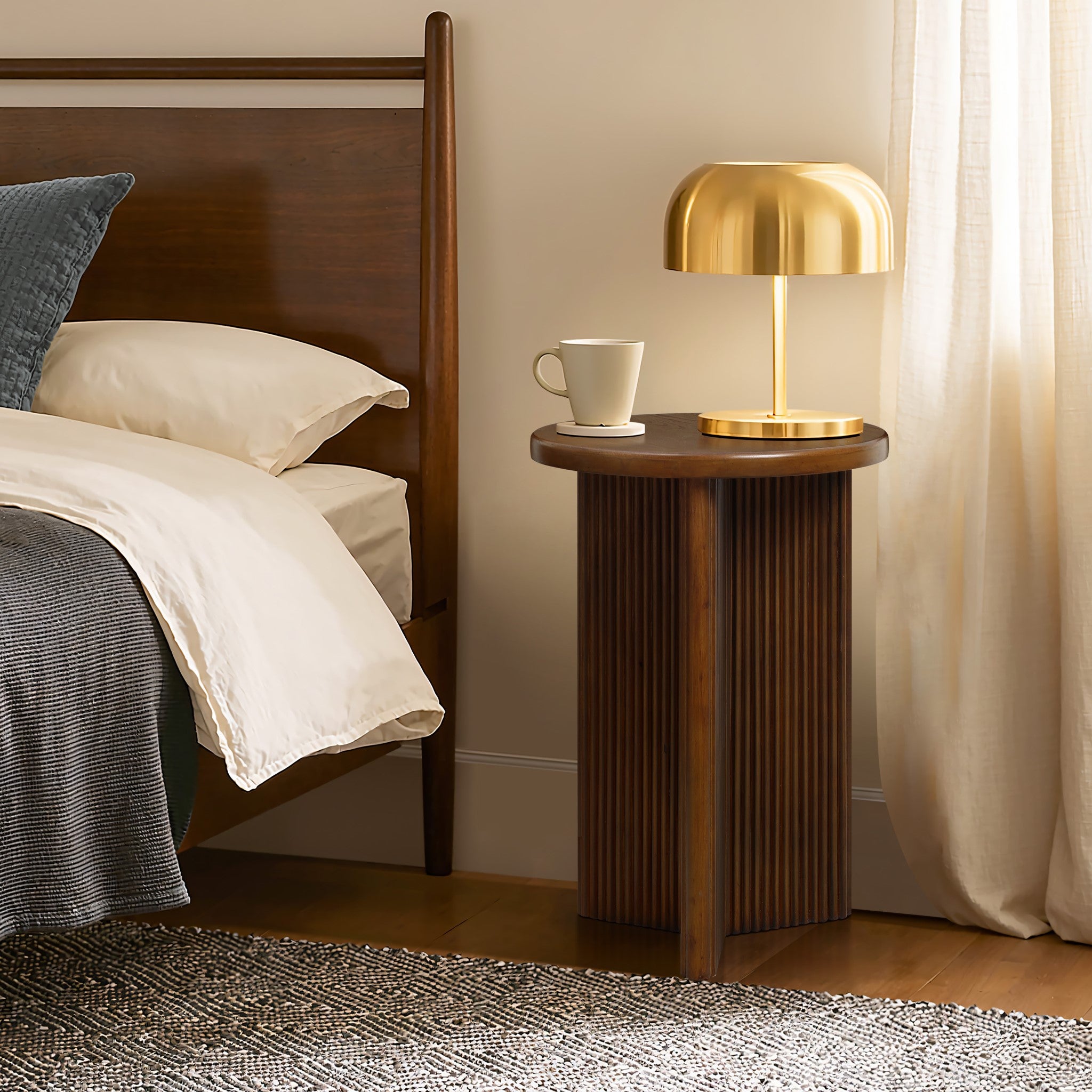 A mid-century modern side table with a round tabletop holds a golden lamp and a white mug beside a bed adorned with white linens. The room features a woven rug and curtains, adding an elegant touch. Crafted from high-quality MDF wood, it exudes both style and durability.
