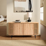 A mid-century modern buffet cabinet with vertical slats, topped with a lamp, books, and a decorative object. Crafted in oak wood grain finish, it sits in a bright living room near a window with a beige rug and part of a sofa visible.