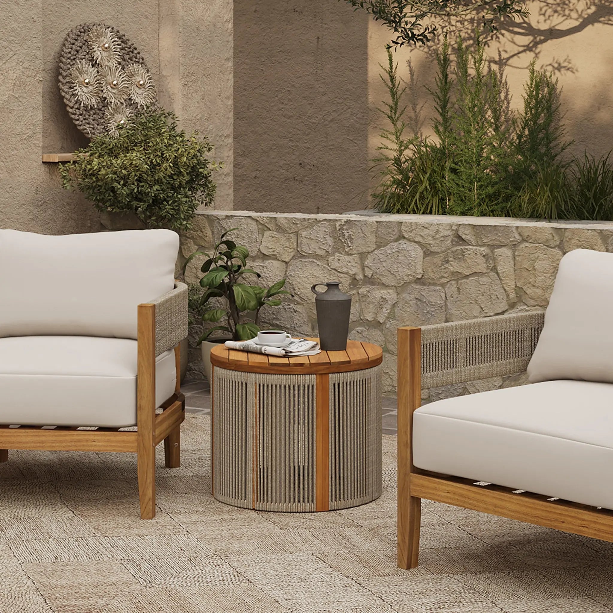A charming outdoor patio scene featuring two wooden armchairs with white cushions, a round drum-shaped side table crafted from natural acacia wood holding a black pitcher and cups. The setting is enhanced by a stone wall backdrop adorned with lush plants.