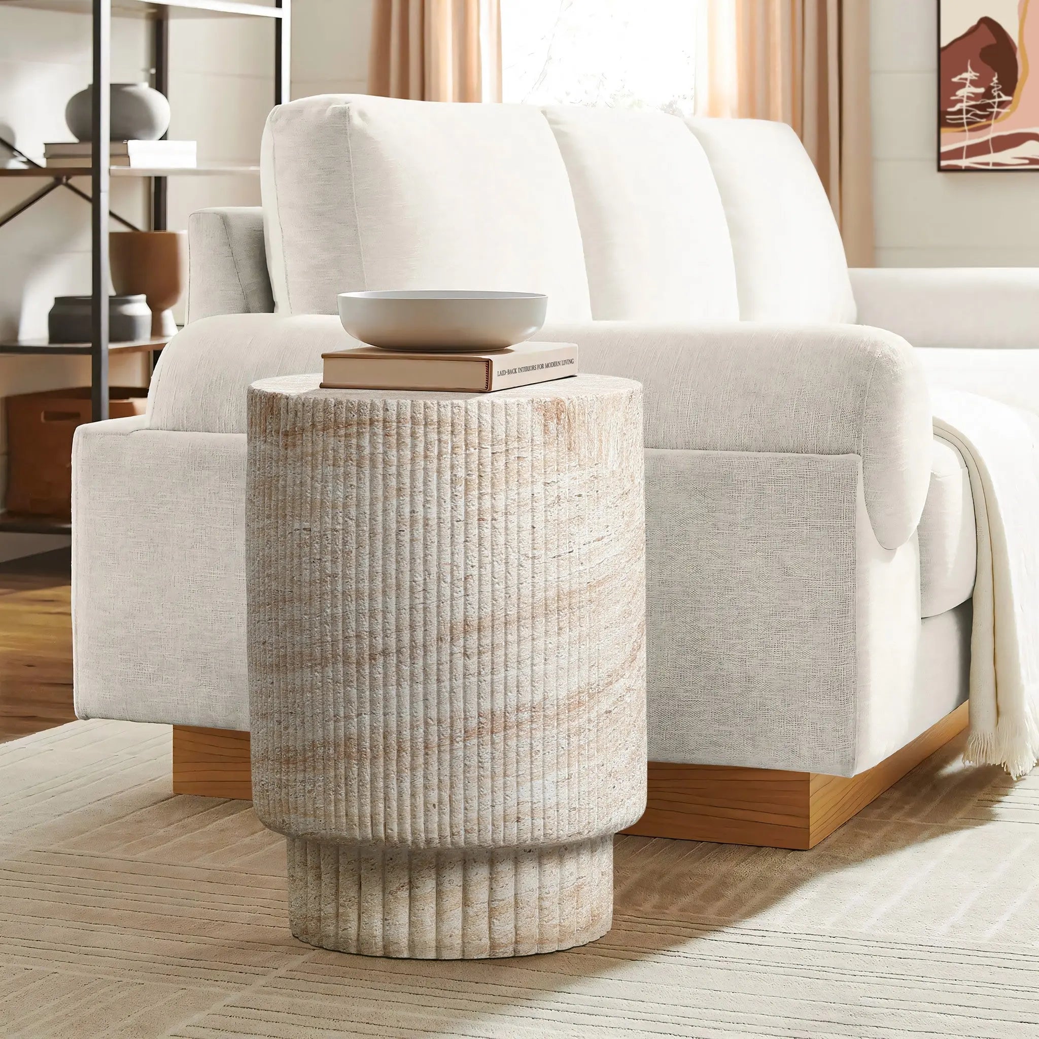 A travertine-style side table crafted from MgO material, holding two books and a bowl, stands beside a white sofa in a living room setting with neutral tones.