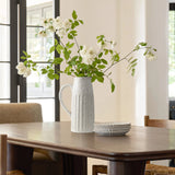 White flowers in a rustic charm ceramic pitcher vase sit on a wooden table, alongside stacked white plates, in a room illuminated by natural light from large windows.