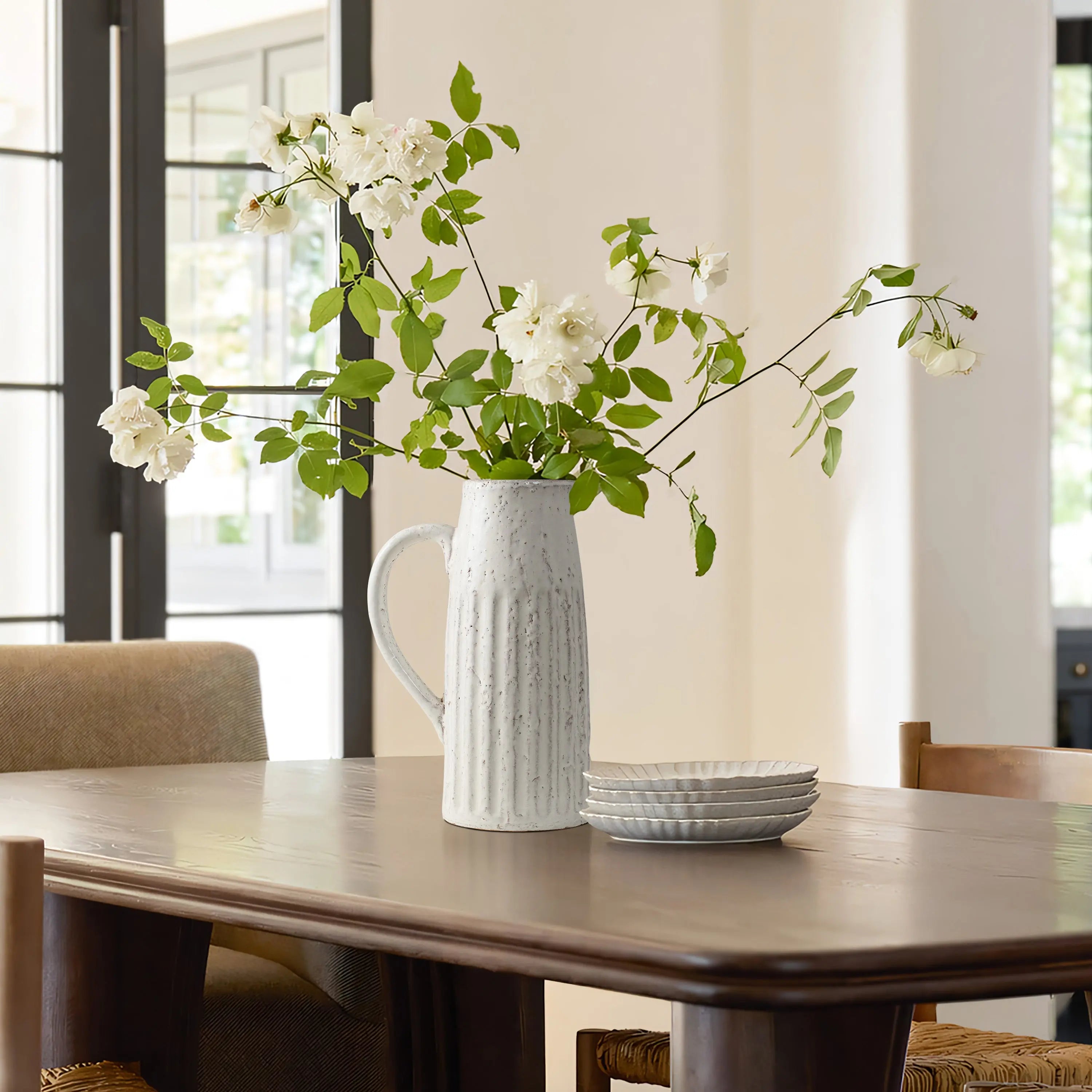 White flowers in a rustic charm ceramic pitcher vase sit on a wooden table, alongside stacked white plates, in a room illuminated by natural light from large windows.