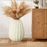 A modern white vase filled with pampas grass stands as an artistic centerpiece on a wooden floor next to a wooden cabinet, against a white paneled wall.