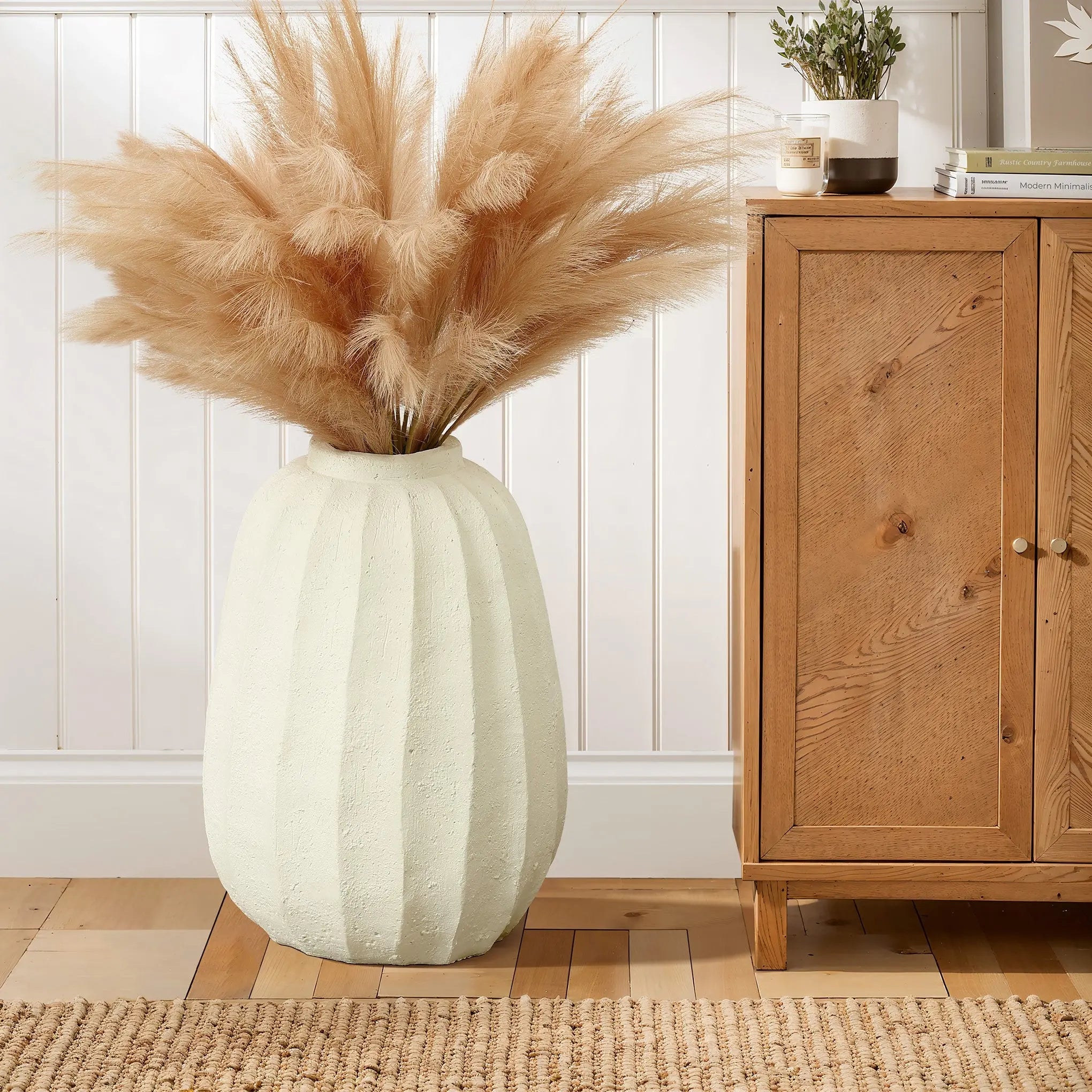 A modern white vase filled with pampas grass stands as an artistic centerpiece on a wooden floor next to a wooden cabinet, against a white paneled wall.