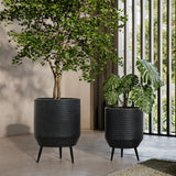 Two tall black planters with green plants stand on a patio. One contains a leafy tree, and the other hosts a large-leafed plant. The pot planter set, in distressed black finish, contrasts beautifully against the vertical slats that provide a backdrop.