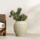 A textured ceramic pot with a green succulent plant sits gracefully on a light-colored floor beside a piece of wooden furniture, showcasing its boho planter charm.