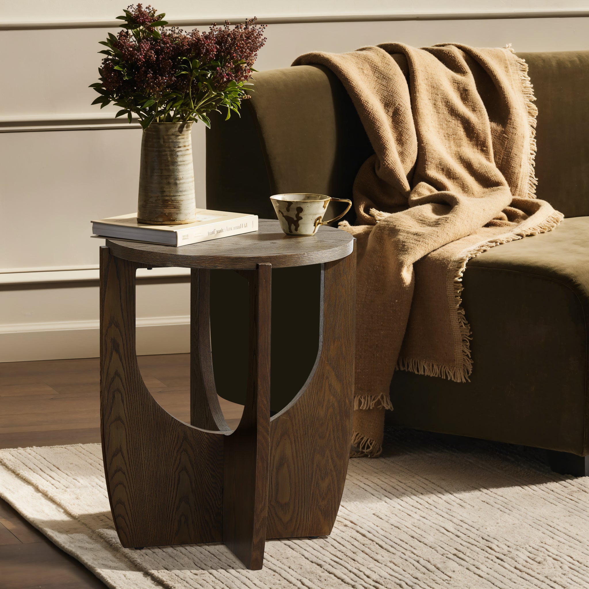 A mid-century modern side table with a walnut veneer holds a vase of flowers, a book, and a teacup. It sits elegantly beside a green sofa adorned with a draped brown blanket.