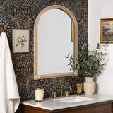 A bathroom with floral wallpaper features a bamboo-framed mirror with rounded edges, gold faucet fixtures, a white countertop, a candle, a potted plant, and framed art on the wall.