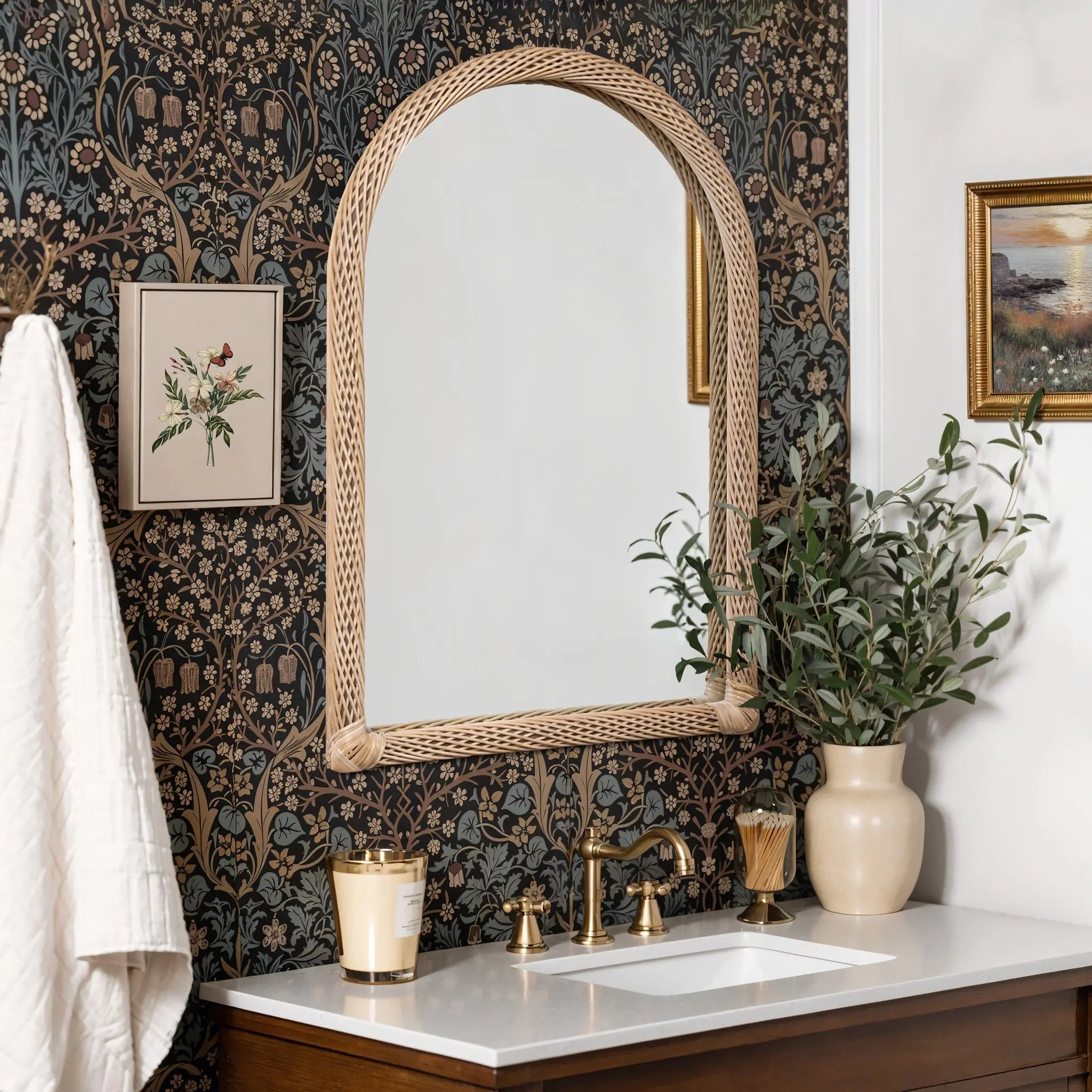 A bathroom with floral wallpaper features a bamboo-framed mirror with rounded edges, gold faucet fixtures, a white countertop, a candle, a potted plant, and framed art on the wall.