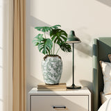 A green leafy plant in a floral ceramic vase sits on a nightstand next to a black desk lamp, books.