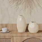 Two ceramic vases with abstract shapes serve as the perfect accent pieces on a wooden cabinet next to an ivory white teacup. A decorative wall hanging graces the background, completing the artistic arrangement.