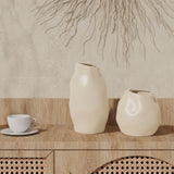 Two ceramic vases with abstract shapes serve as the perfect accent pieces on a wooden cabinet next to an ivory white teacup. A decorative wall hanging graces the background, completing the artistic arrangement.