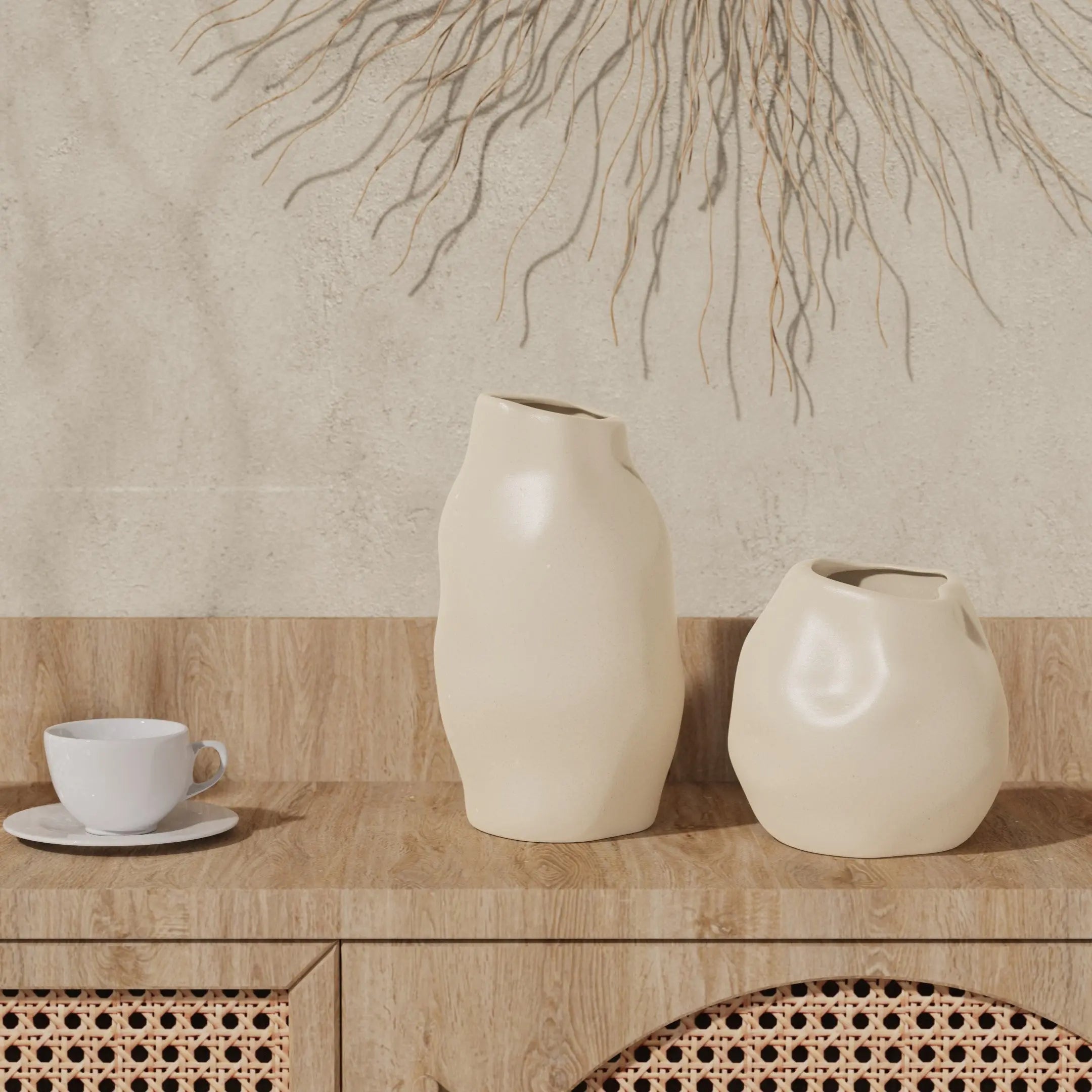 Two ceramic vases with abstract shapes serve as the perfect accent pieces on a wooden cabinet next to an ivory white teacup. A decorative wall hanging graces the background, completing the artistic arrangement.