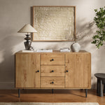 Wood buffet cabinet with three drawers two cabinets stands against a beige wall, topped with a lamp, books, and a vase.