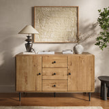 Wood buffet cabinet with three drawers two cabinets stands against a beige wall, topped with a lamp, books, and a vase.