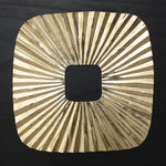 Square gold metallic object with a central square cutout and radiating, textured lines evokes striking sunburst wall art.