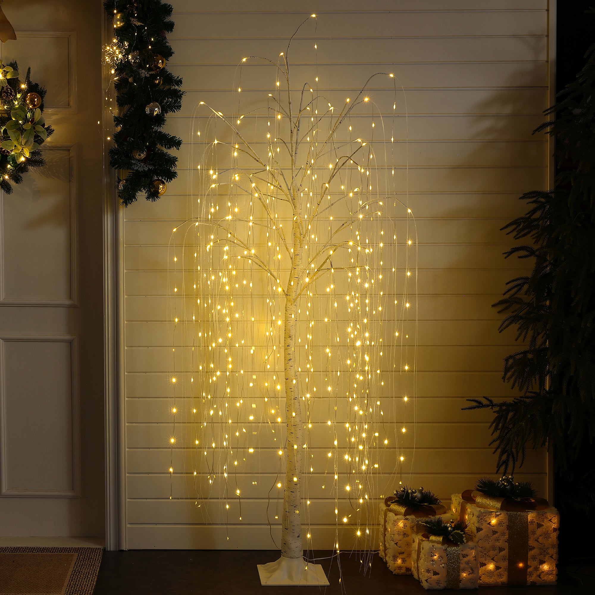 Artificial willow christmas Tree with cascading warm white string lights stands indoors near wall, wrapped presents greenery.