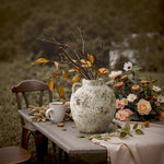 Terracotta Vase with red maple branches sits on a round mat beside a wooden chair draped with a beige blanket on a porch, surrounded by autumn foliage—perfect for cozy rustic home décor.