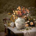 Terracotta Vase with red maple branches sits on a round mat beside a wooden chair draped with a beige blanket on a porch, surrounded by autumn foliage—perfect for cozy rustic home décor.