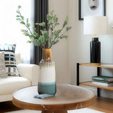 A colorful vase with leafy branches sits on a round wooden table in the living room, surrounded by home decor items like a couch, decorative pillows, and a shelf lamp in the background.