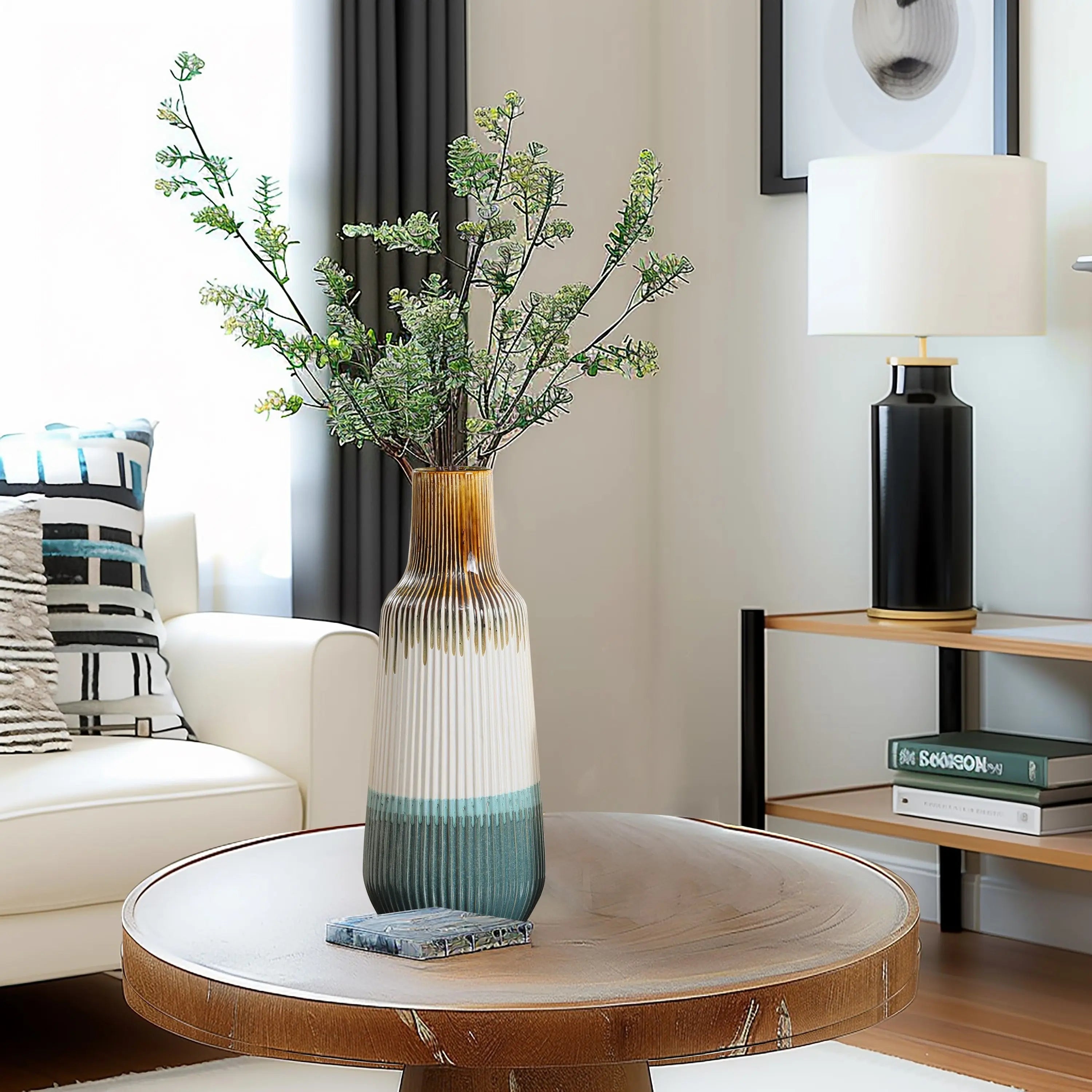A colorful vase with leafy branches sits on a round wooden table in the living room, surrounded by home decor items like a couch, decorative pillows, and a shelf lamp in the background.