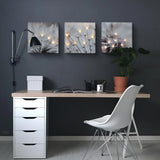 Minimalist desk setup with a white chair, black lamp, potted plant, open notebook, and three glowing canvas art dandelion prints beautifully illuminated on a dark gray wall.