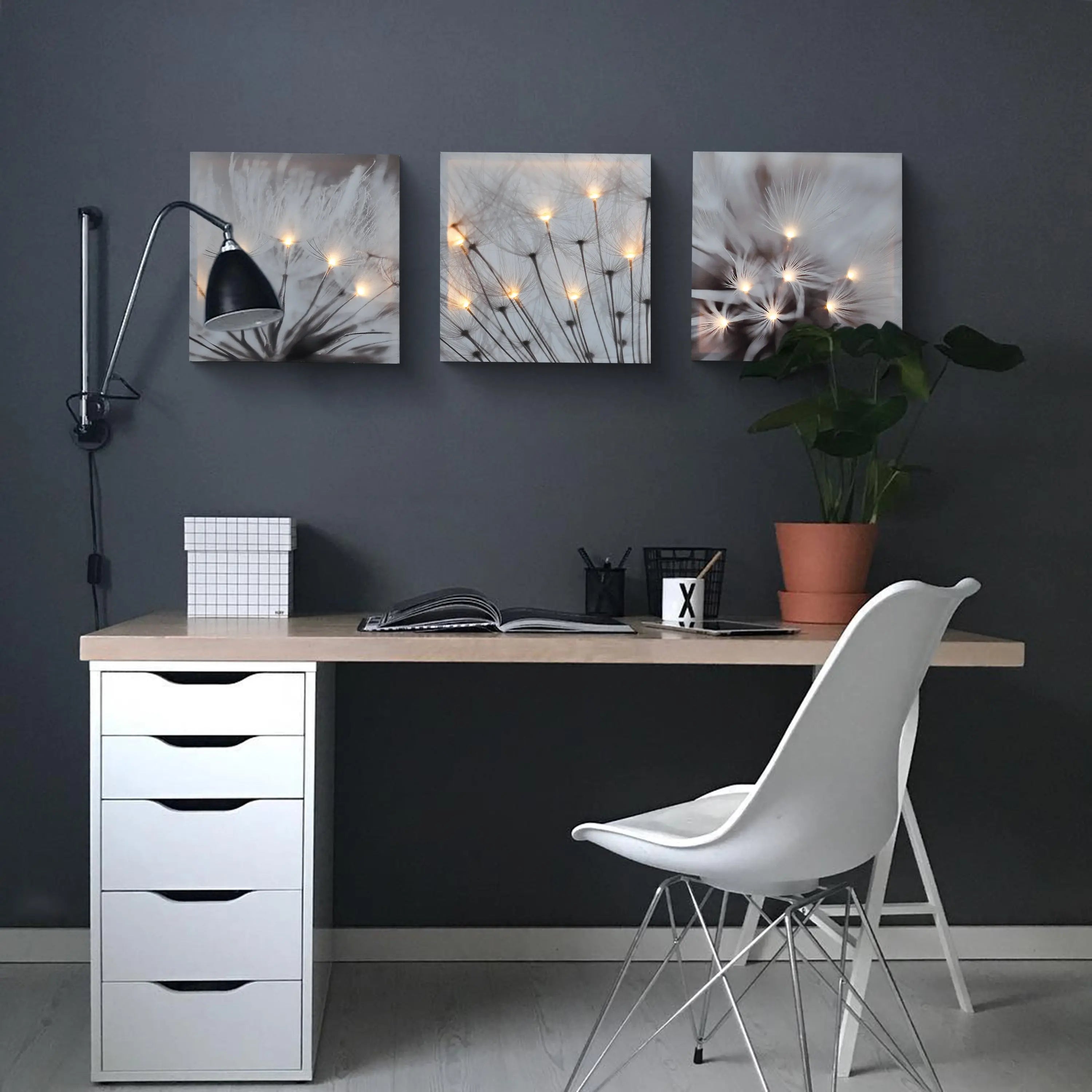 Minimalist desk setup with a white chair, black lamp, potted plant, open notebook, and three glowing canvas art dandelion prints beautifully illuminated on a dark gray wall.