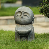 A small Buddha monk statue holding a bowl, sitting on grass with bushes and a stone pathway in the background, makes a charming garden statue or meditation centerpiece.