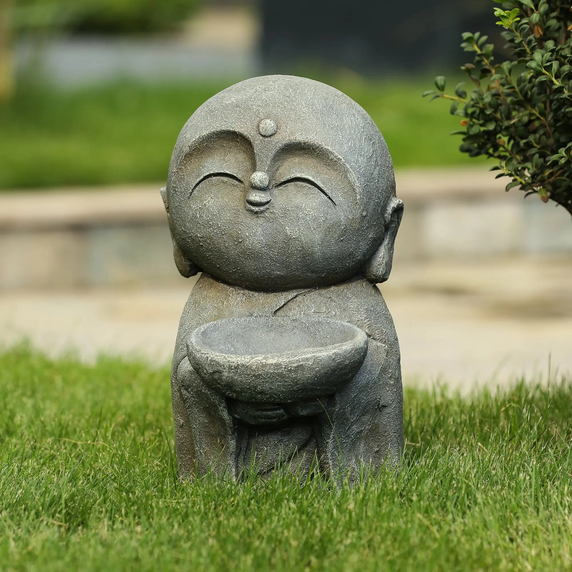 A small Buddha monk statue holding a bowl, sitting on grass with bushes and a stone pathway in the background, makes a charming garden statue or meditation centerpiece.