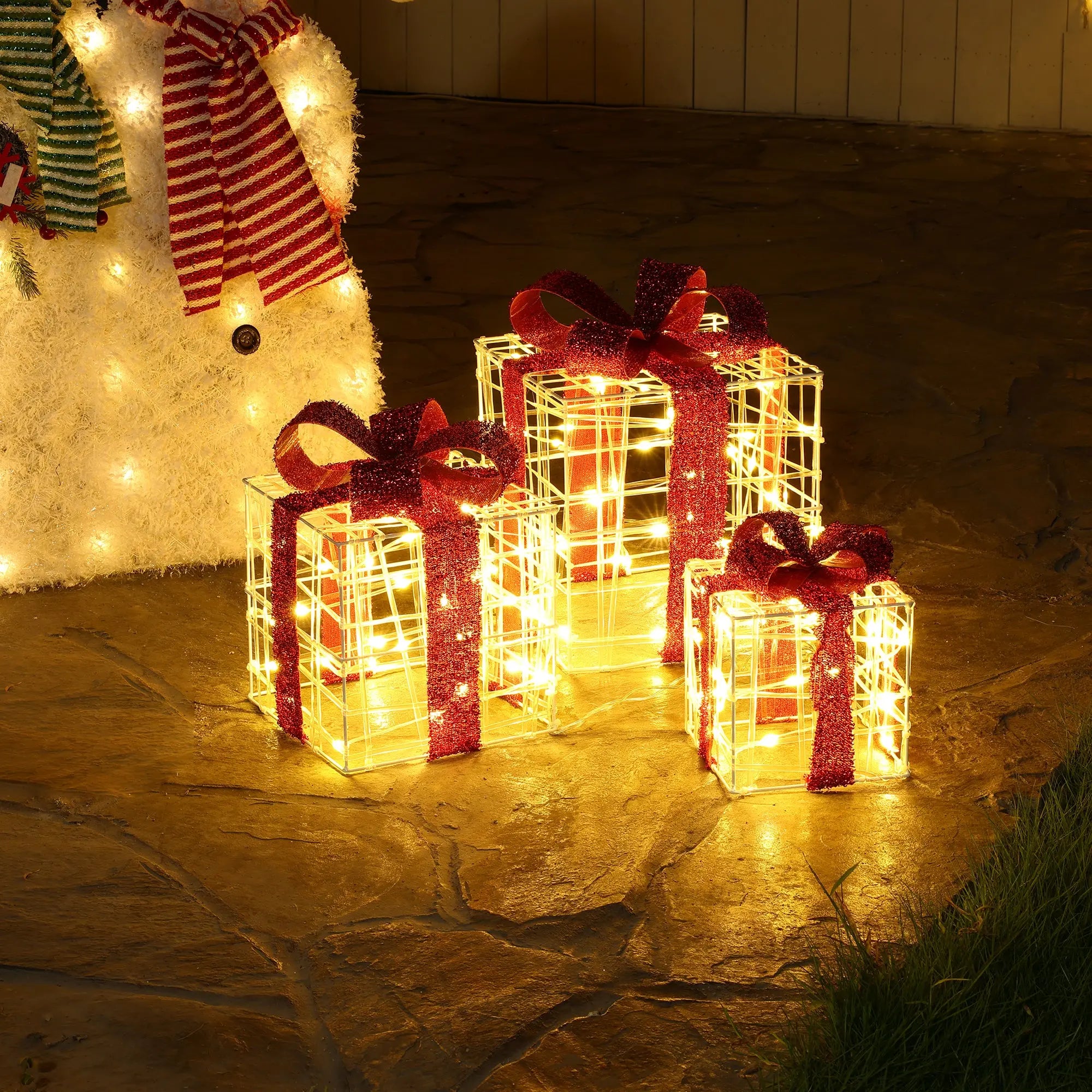 Three lighted gift boxes with red bows are displayed on a stone patio next to a lit snowman figure, creating charming holiday decorations.