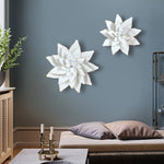 Two white lotus wall decorations, reminiscent of metallic art, elegantly adorn a gray wall above a sofa in a living room adorned with plants and candles on the table.